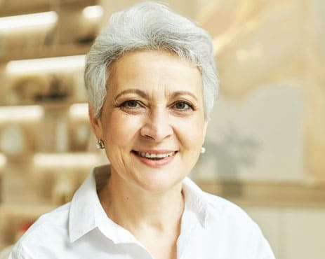 Smiling mature woman with gray hair wearing a white shirt in a dental office setting.