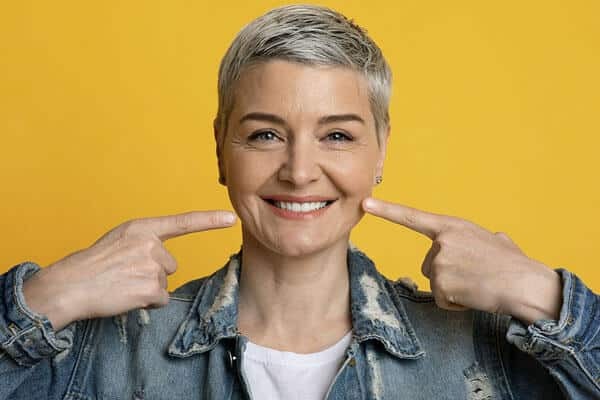 Woman smiling while pointing to her teeth against a yellow background.