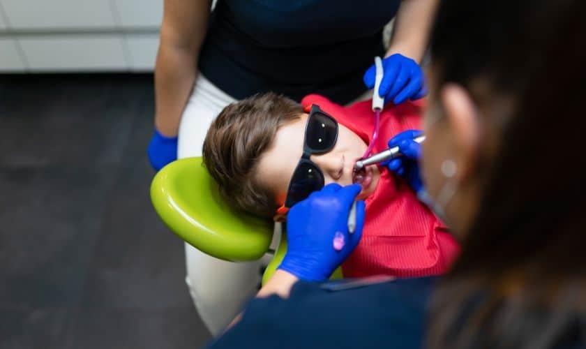 Child wearing sunglasses and red and blue clothing holds a green toy while receiving preventive dental care.