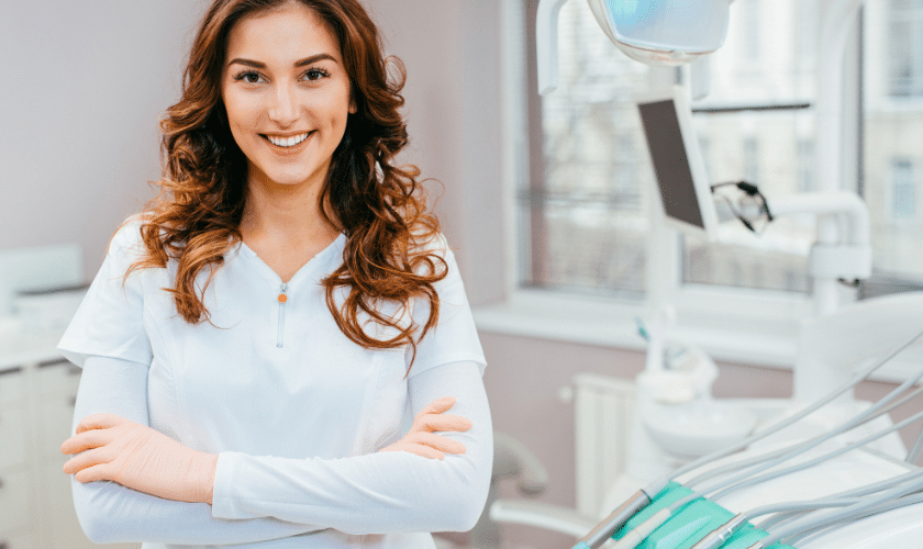 Smiling female dentist in white uniform standing in modern dental office treatment room.