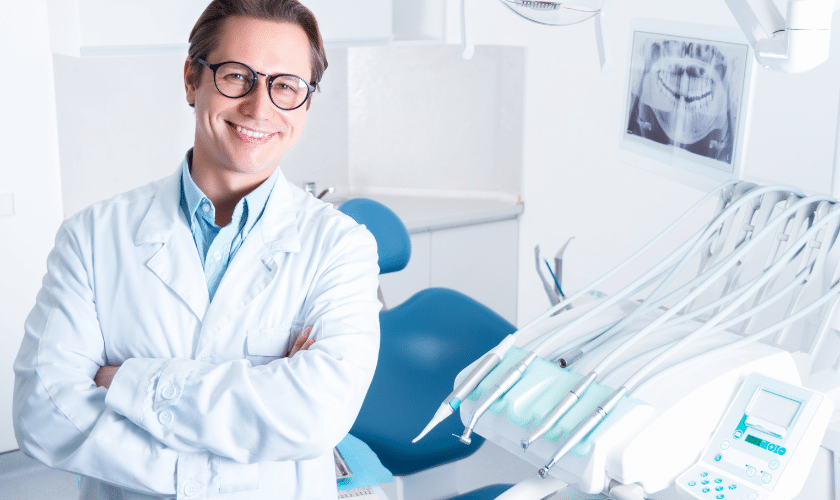 Smiling male dentist in white coat and glasses standing in modern dental office with blue chair.