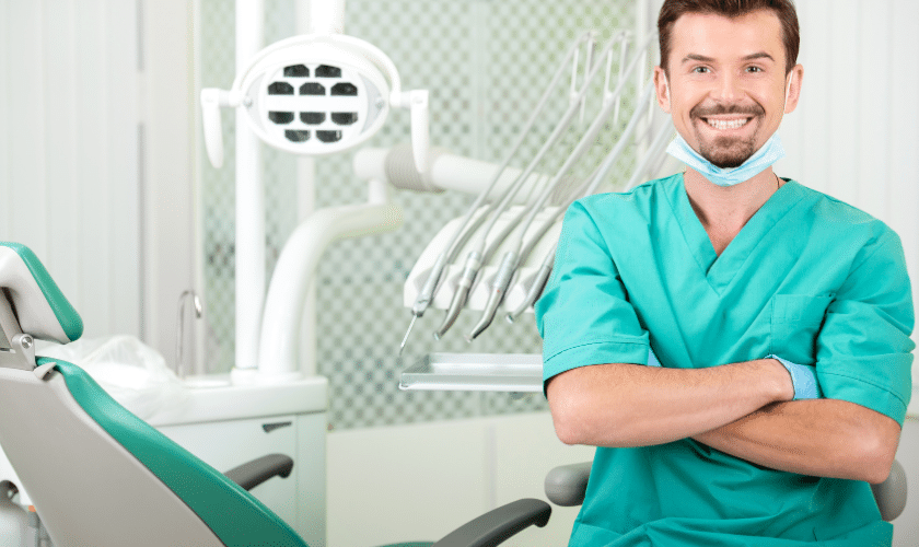 Smiling dentist in turquoise scrubs in modern dental operatory with chair and overhead light.