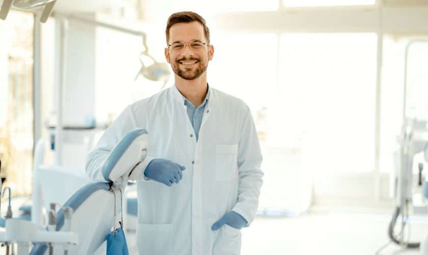 Smiling dentist in white coat with blue gloves standing in modern dental office.