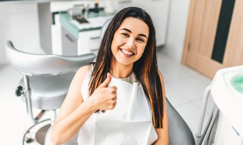 Happy young woman giving thumbs up in modern dental office treatment room.