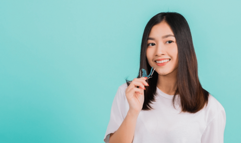 Young woman holding an Invisalign aligner, smiling against a turquoise background.