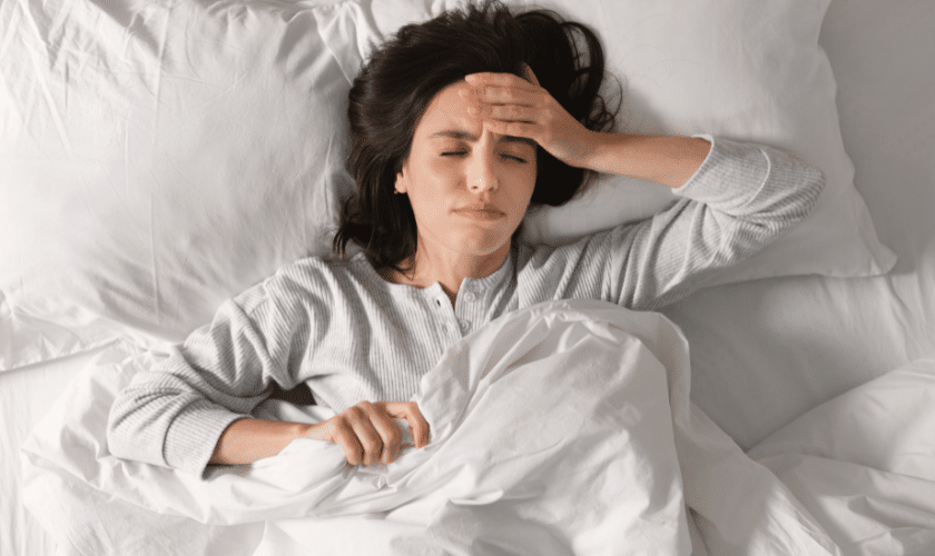 Woman in bed experiencing sleep apnea symptoms, holding her forehead in discomfort.