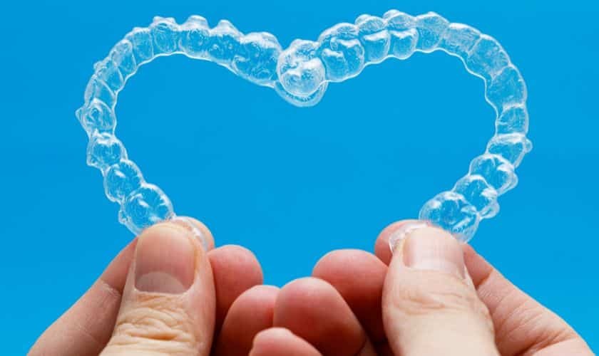 Hands holding clear Invisalign aligners shaped like a heart against blue background.