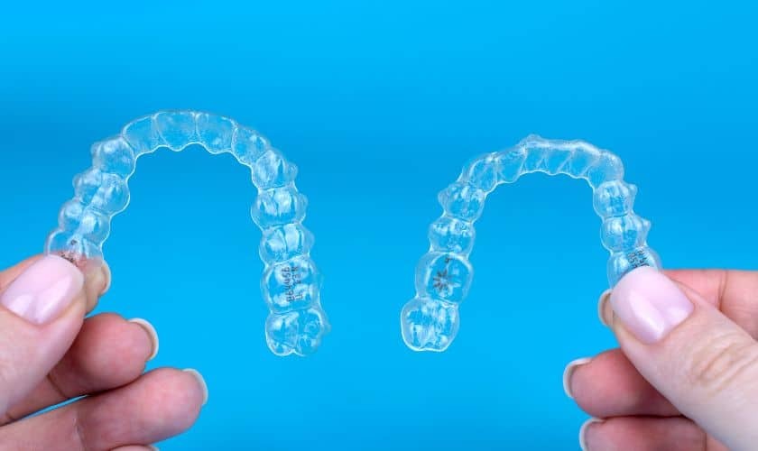 Hands holding two clear Invisalign aligners against a blue background.