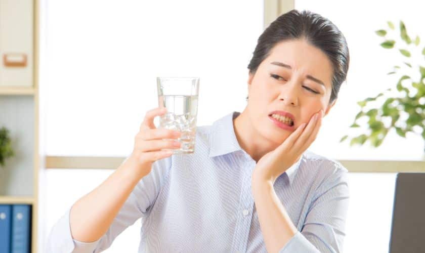 Woman holding glass of water while touching her cheek, experiencing tooth sensitivity pain.