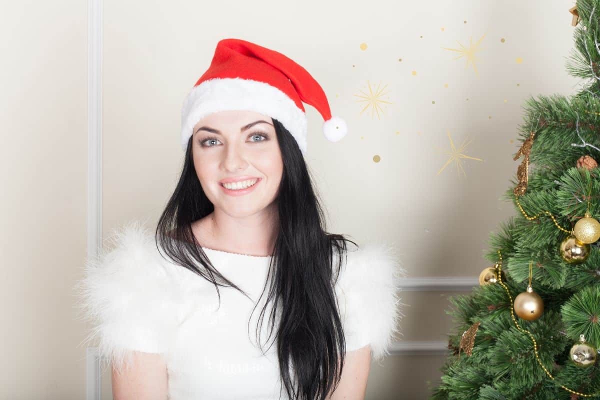 Woman wearing Santa hat smiling brightly next to decorated Christmas tree.