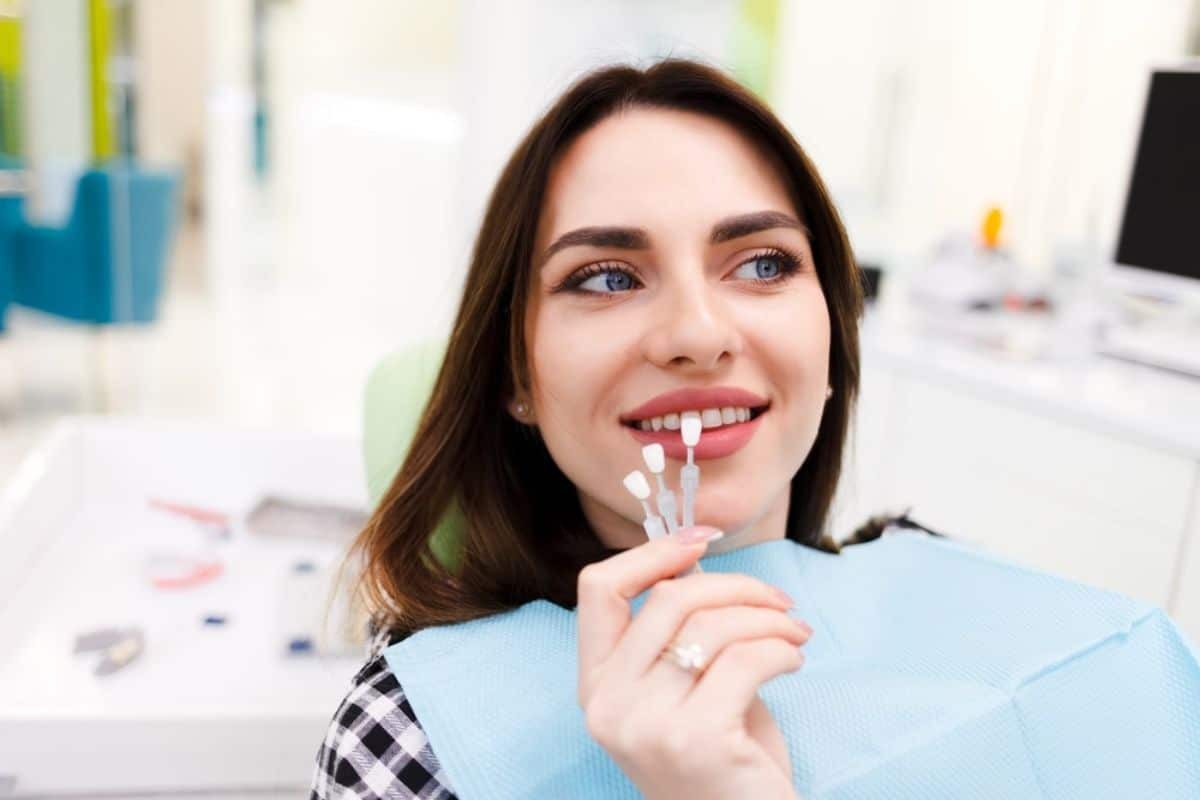 Young woman smiling while holding porcelain veneers in a dental office setting.