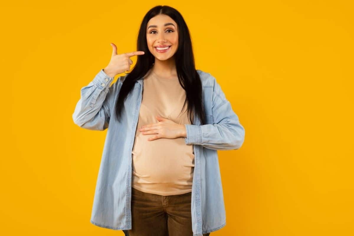 Pregnant woman pointing to her smile against a yellow background.