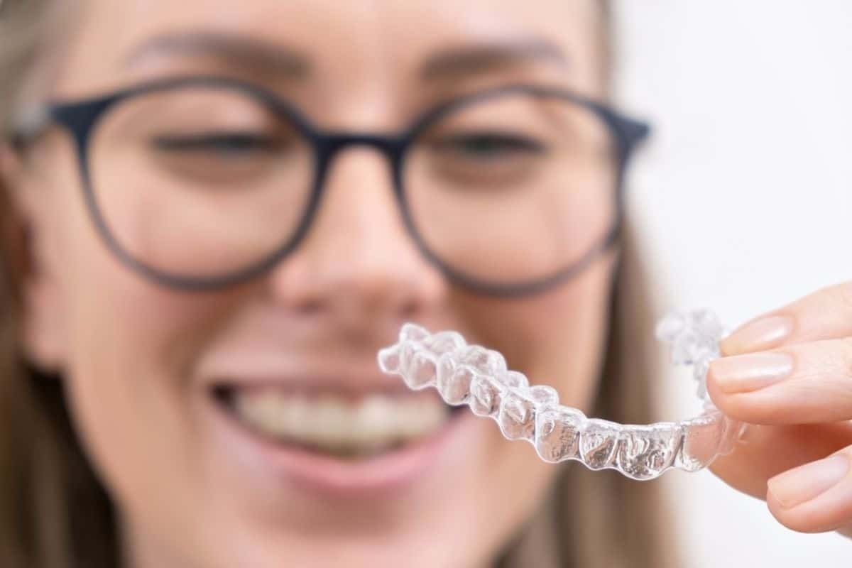 Smiling woman wearing glasses holds a clear aligner tray for Invisalign treatment.
