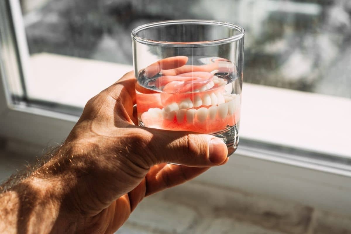 Hand holding a glass of water with dentures submerged in red cleaning solution.