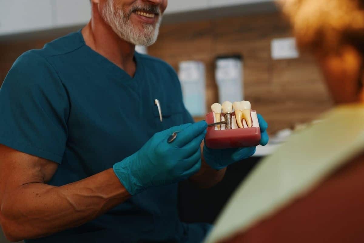 Dentist in blue scrubs demonstrates dental implant model to patient during consultation.