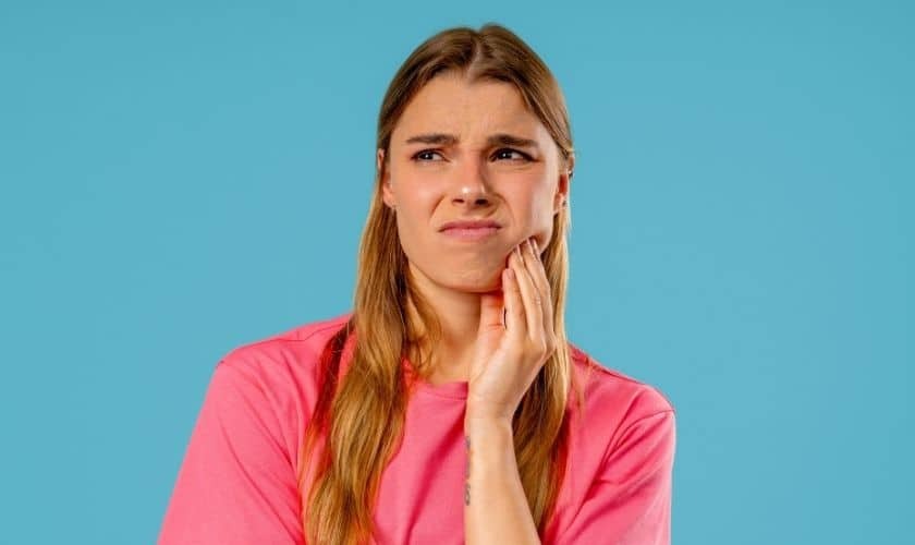 Woman with tooth pain holding her cheek against blue background for dental emergency article.
